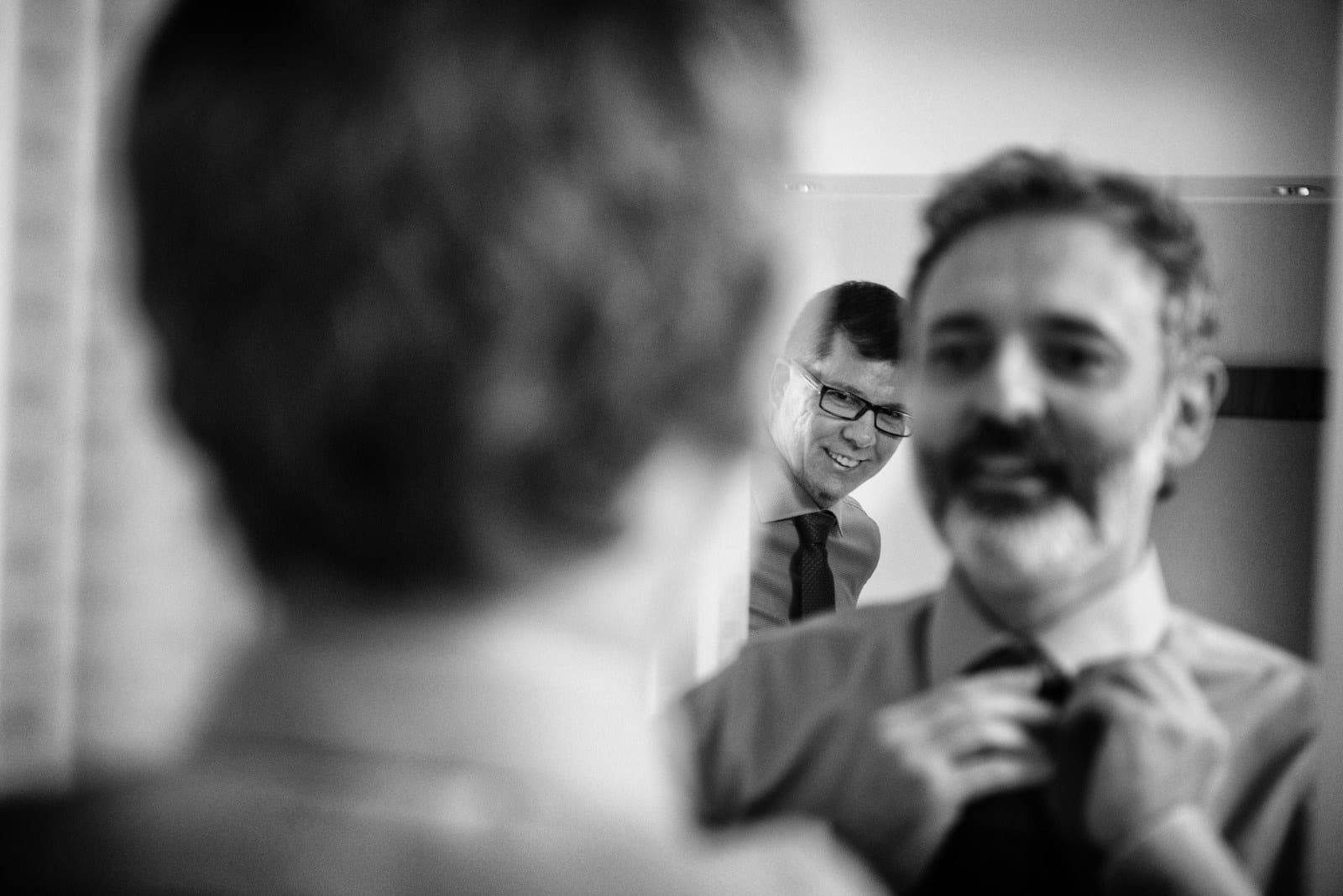 A bearded man adjusts his tie in a mirror as his husband-to-be looks from behind.