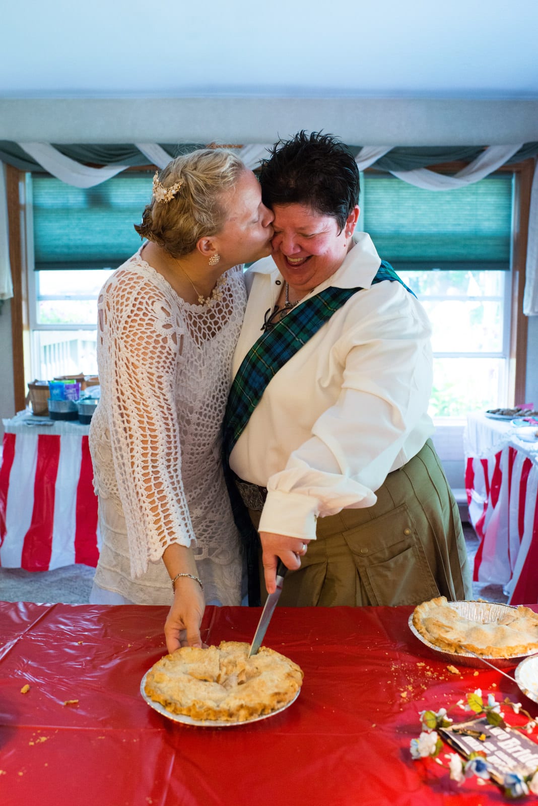 Two brides cut a pie during their wedding. One of the brides kisses the cheek of the other.