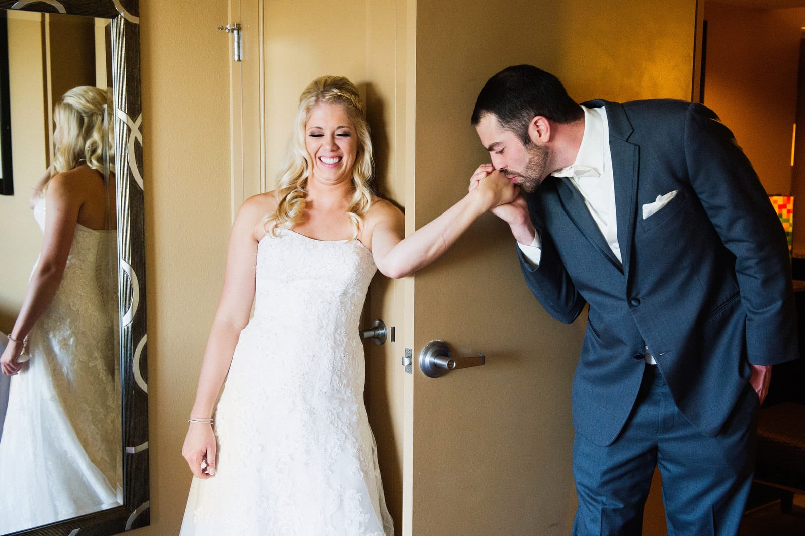 A bride smiles as her husband-to-be kisses her hand from around the corner of a door because they didn't want to see each other before their wedding at the Embassy Suites near the airport before their wedding.