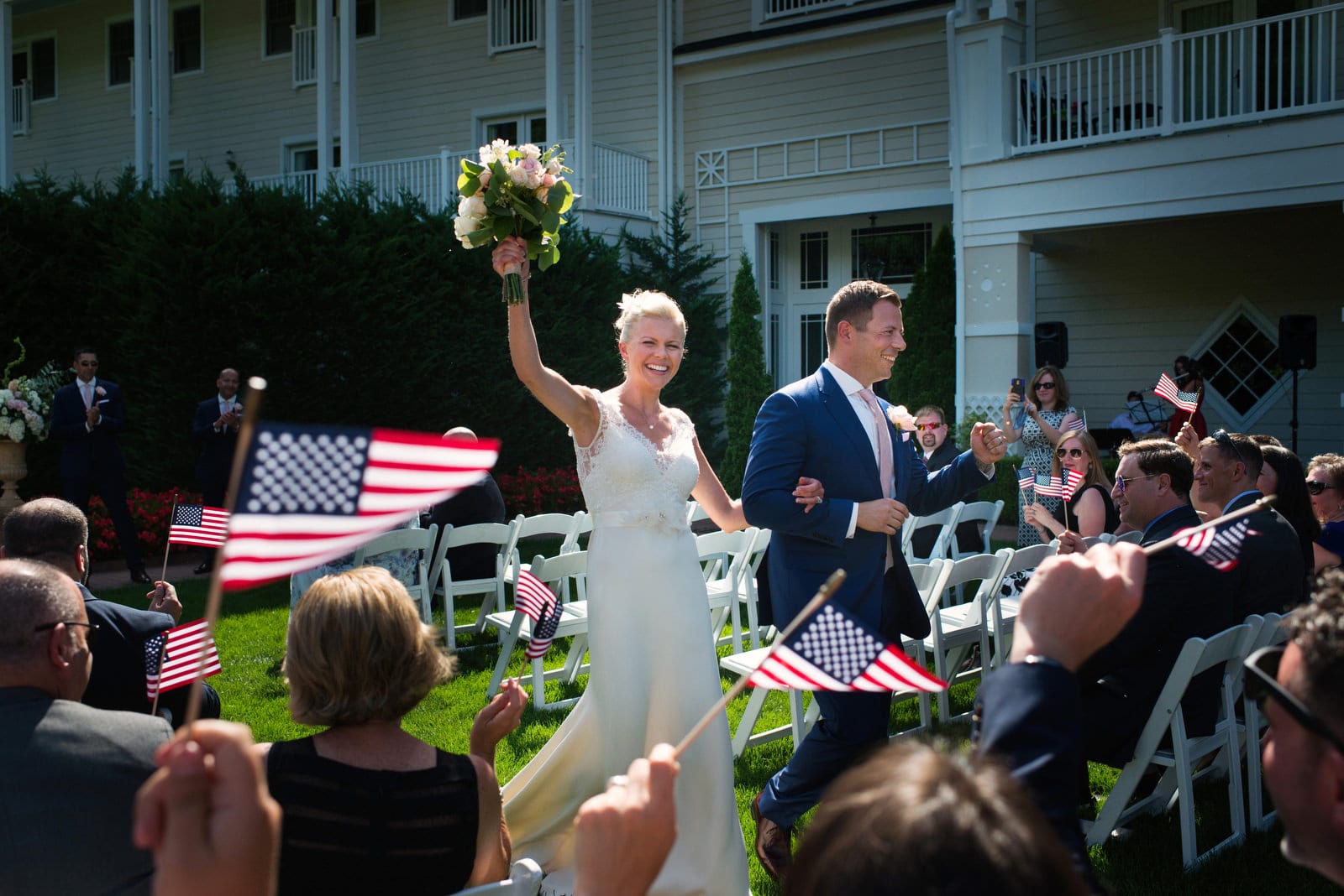 A bride smiles and lifts her bouquet in the air as she and her husband walk down the aisle after their outdoor wedding at the Omni Bedford Springs resort. Their guests are waving American flags because the wedding is on the fourth of July.