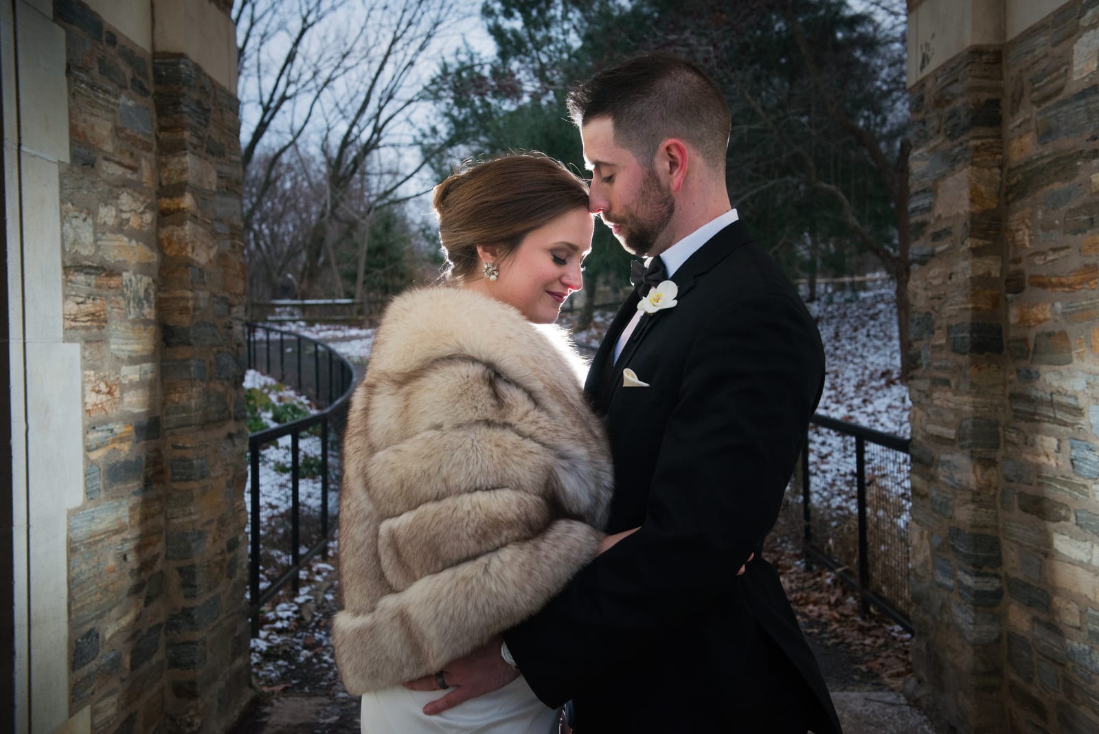 A bride and groom embrace in a stone archway during a chilly winter evening. The bride is wearing a fur stole.