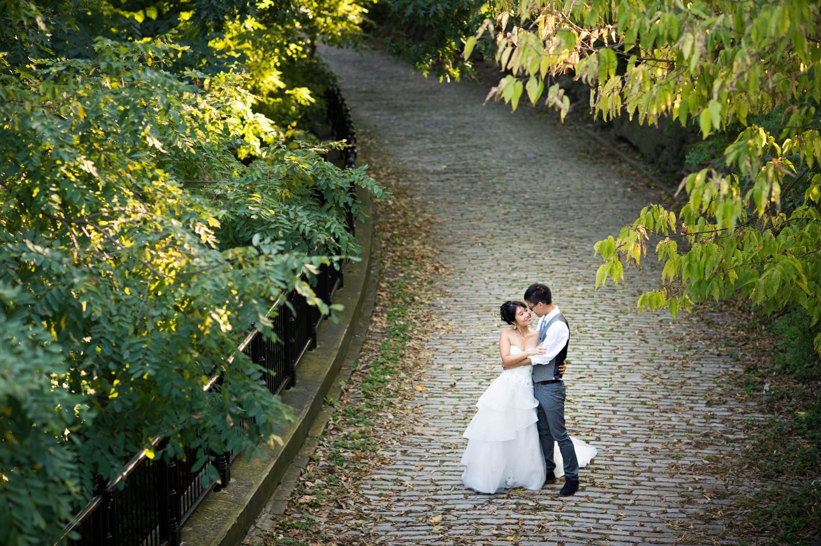 A bride and groom embrace on the paving stone path behind the Schenley Park visitor's center as seen from above.
