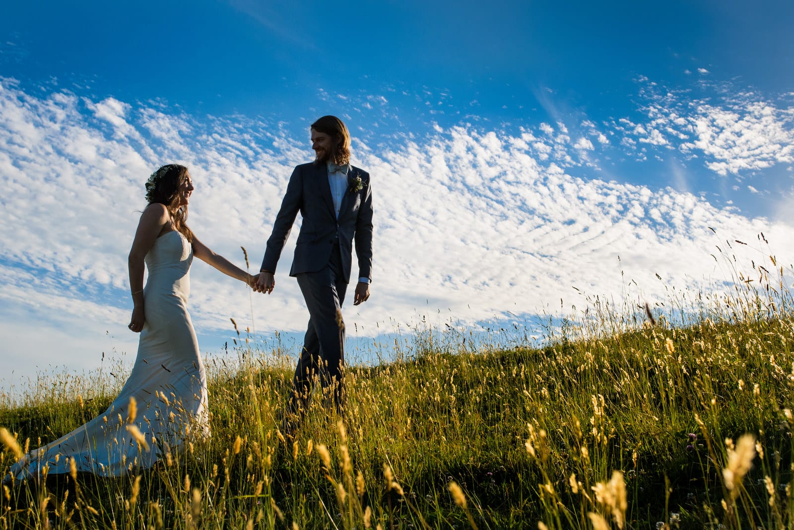 A groom looks back as he leads his bride through a field of tall grass in front of a sky filled with wispy clouds.