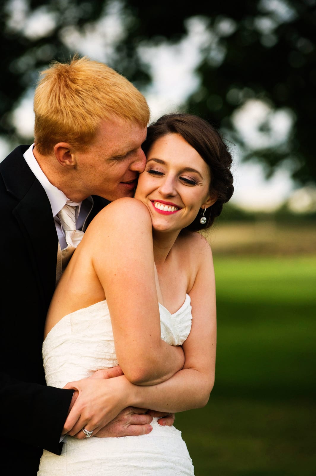 A bride smiles and looks back at her red-headed groom as he holds her from behind and kisses her on the cheek during their wedding at the Montour Heights Country Club.