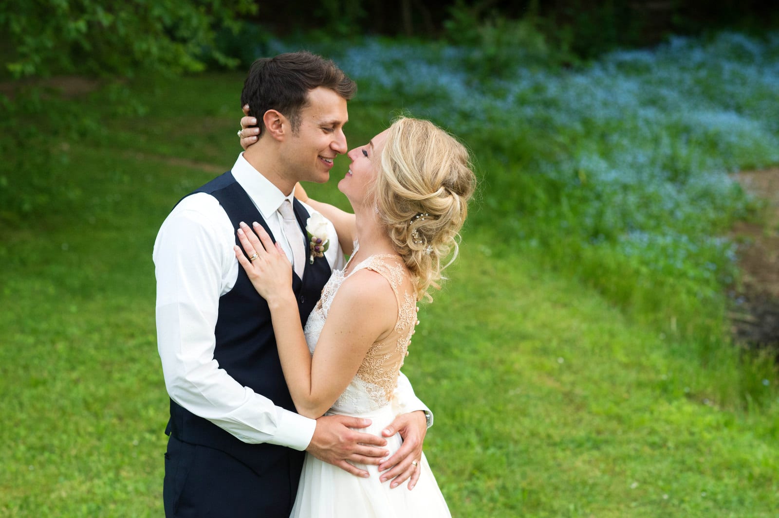 A bride and groom stand face to face and embrace in a field with light blue flowers after their Seven Springs wedding.