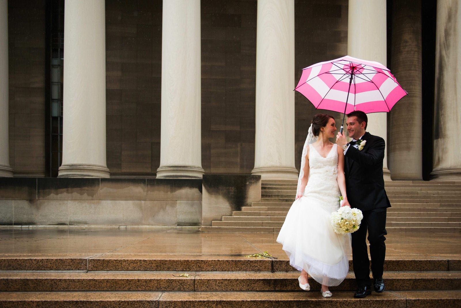 A bride and groom walk down the steps of the Mellon Institute in the rain. He holds a pink and white umbrella over them.