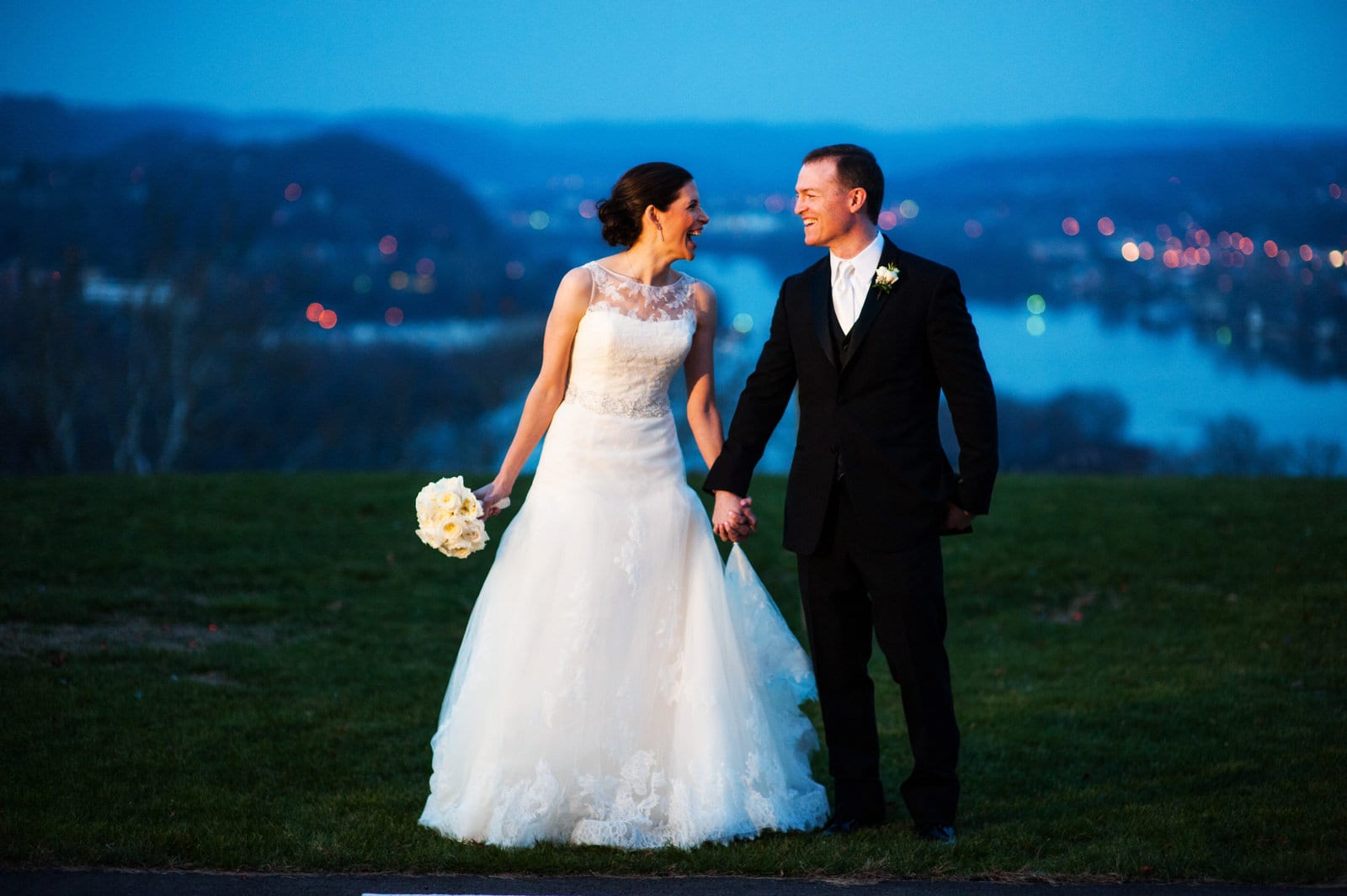 A bride and groom hold hands and look at each other as they stand on a hill high above the Allegheny River at twilight after their wedding at Longue Vue country club.