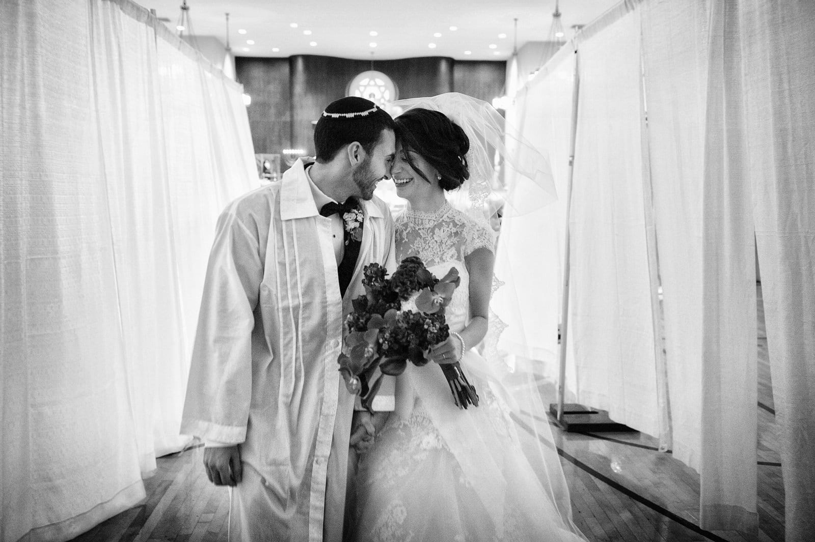 A bride and groom smile and touch foreheads as they walk between curtains put up in the synagogue in Charleston West Virginia.