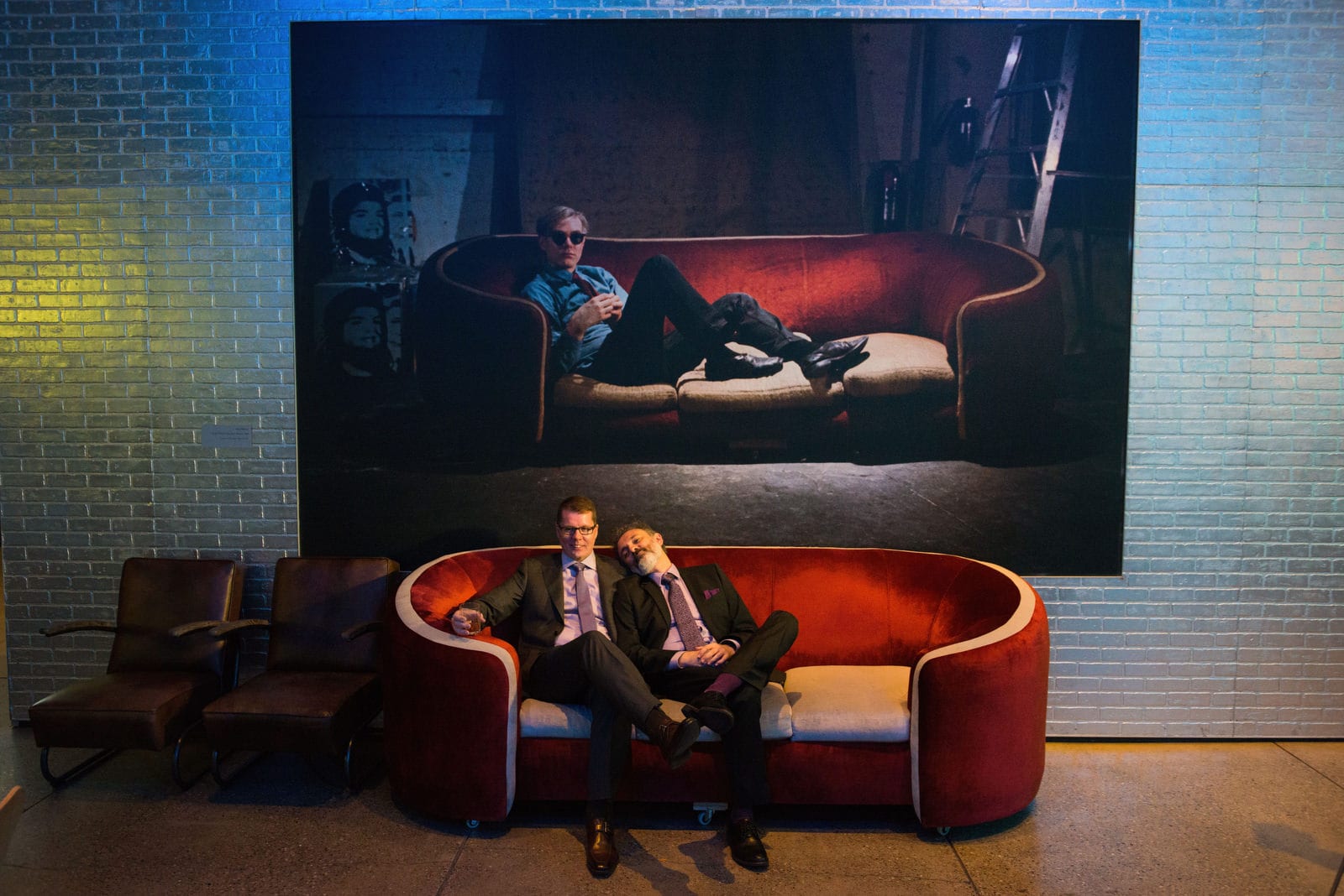 Two grooms recline on a couch after their wedding at the Andy Warhol museum. There is a portrait of Andy Warhol above them sitting on the exact same sofa.
