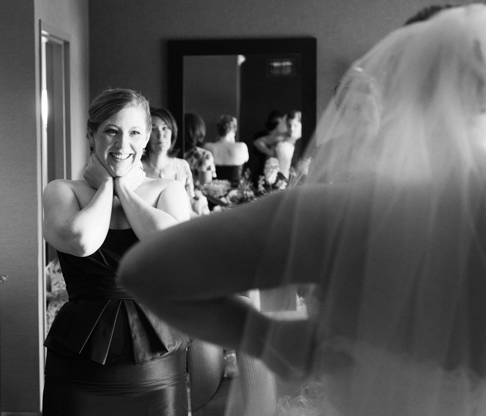 A bridesmaid smiles and holds her hands to her face as she looks at the bride.