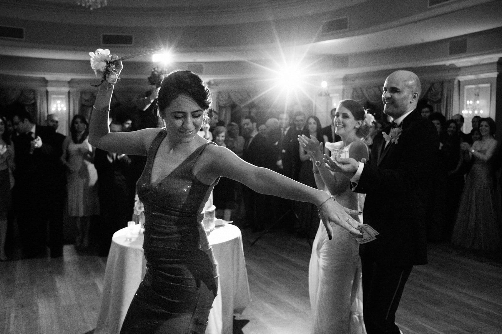 A woman dances with a knife and a ten dollar bill at a Persian wedding at Oheka Castle. The bride and groom smile and applaud behind her.