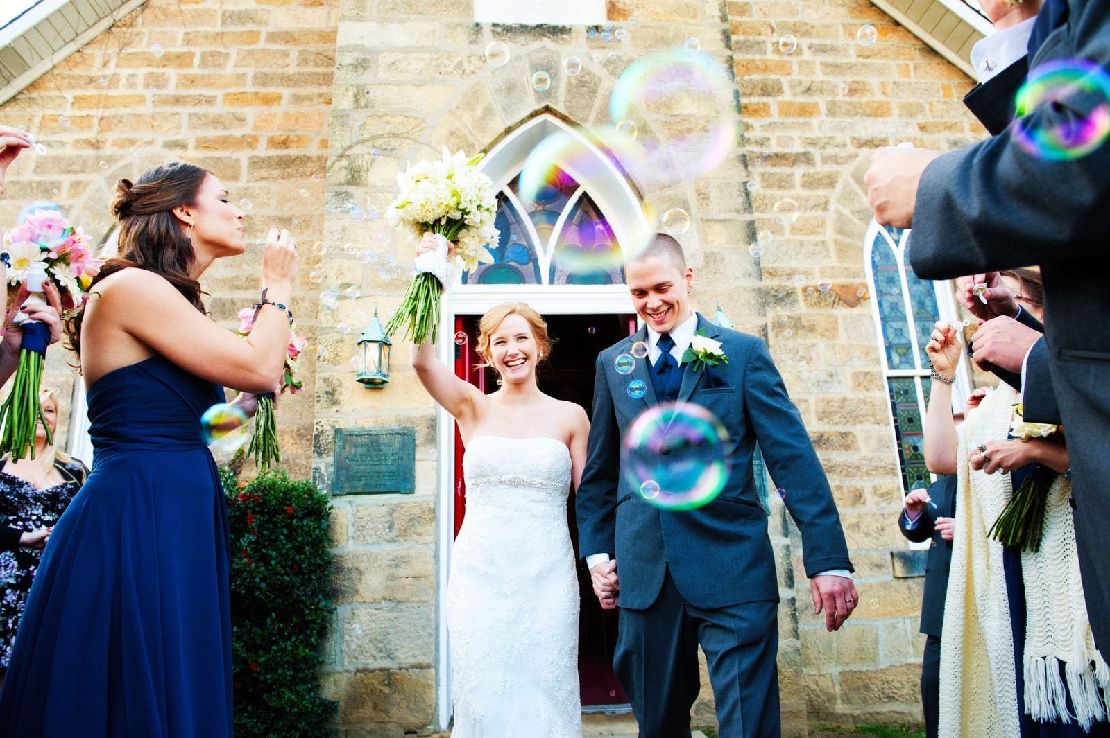 Seen through a cloud of bubbles, a bride and groom exit a stone church after their wedding.