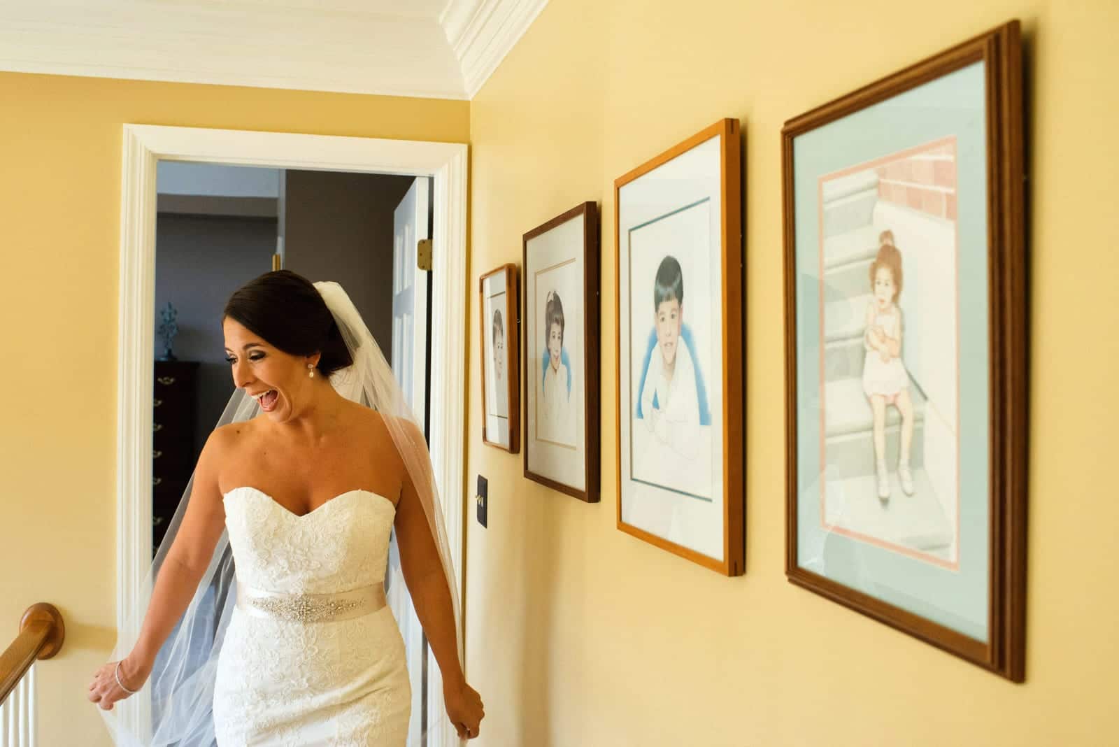 A bride looks down and smiles as she walks on a landing at her parents house with portraits of her and her siblings on the wall behind her.