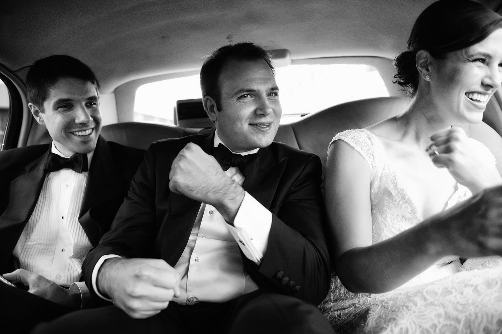 A groom smiles and holds his fist up while seated in the back of a limousine with his bride and her brother before their wedding at the Pittsburgh Center for the Arts.