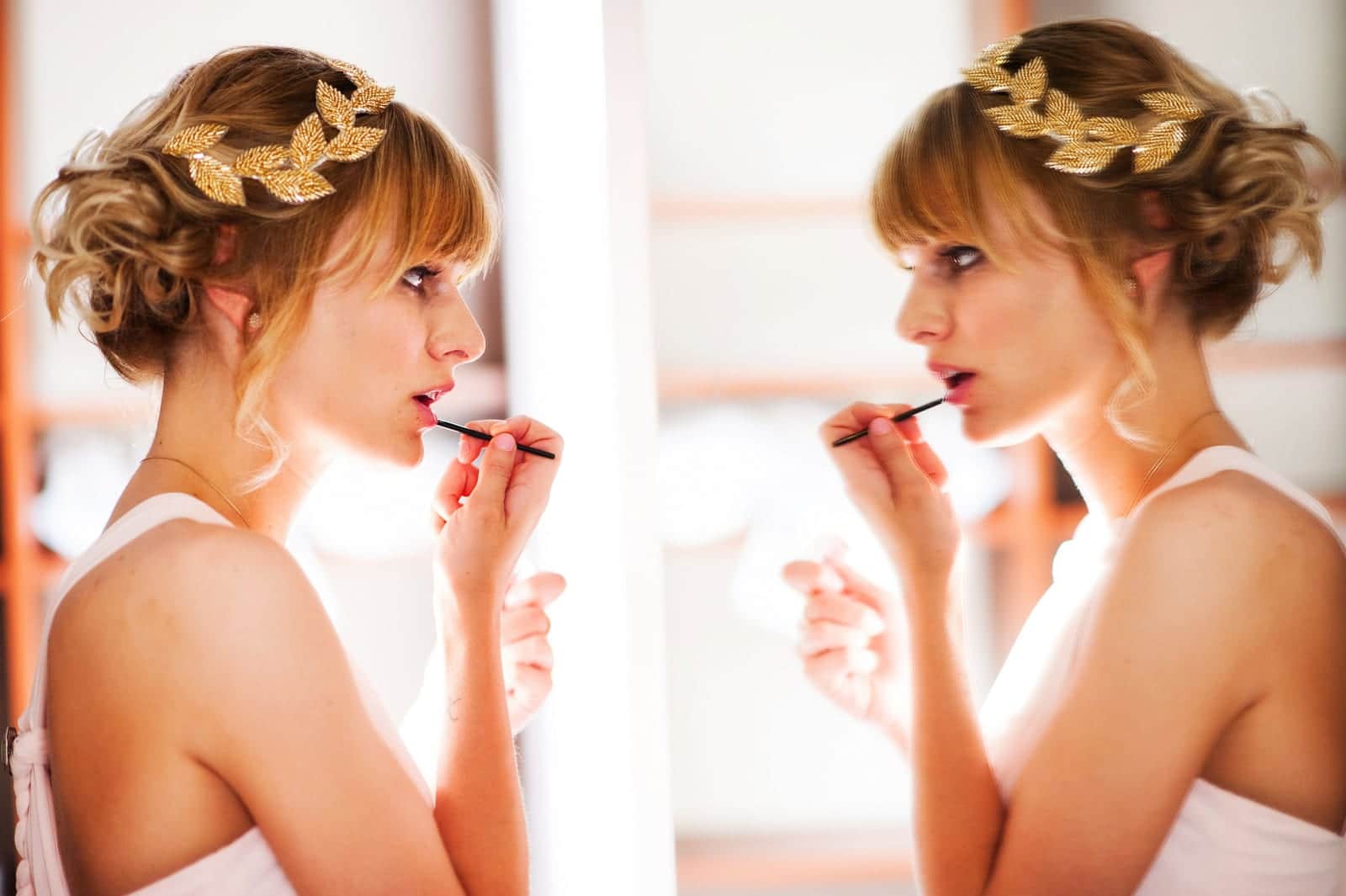 A woman with a gold garland in her hair is reflected in a mirror as she applies lipstick during a wedding at the Mansion at Maple Heights.