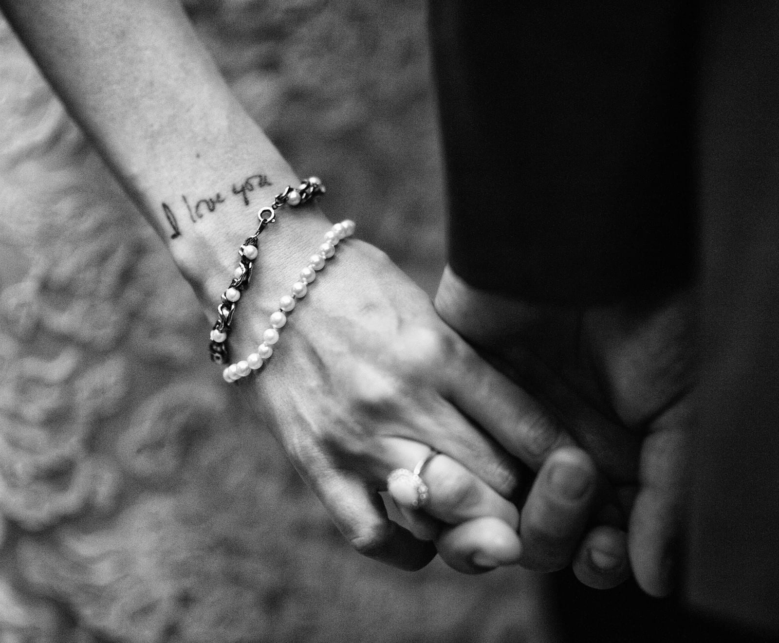A closeup of a bride and groom holding hands shows a tattoo on her wrist that says "I love you"