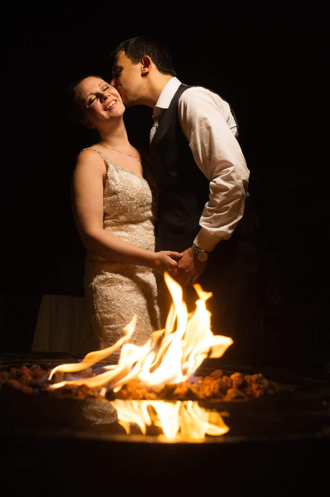 A bride smiles as she stands by a fire pit and her groom kisses her on the cheek after their Seven Springs Wedding.