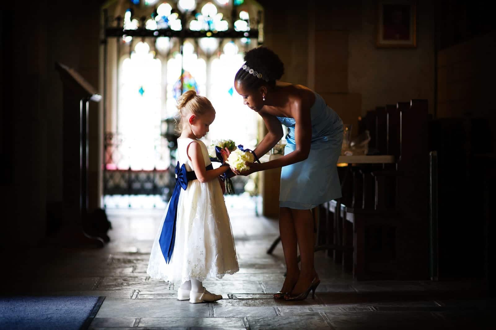 A bridesmaid in a light blue dress helps a flower girl with her bouquet in the back of Sacred Heart Church in Shadyside. The flower girl is wearing a white dress with a blue bow.