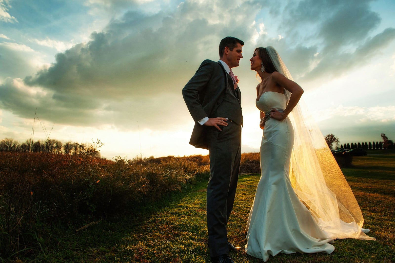 A bride and groom stand facing each other with their arms on their hips and the sun setting behind them during their Lingrow Farm wedding.
