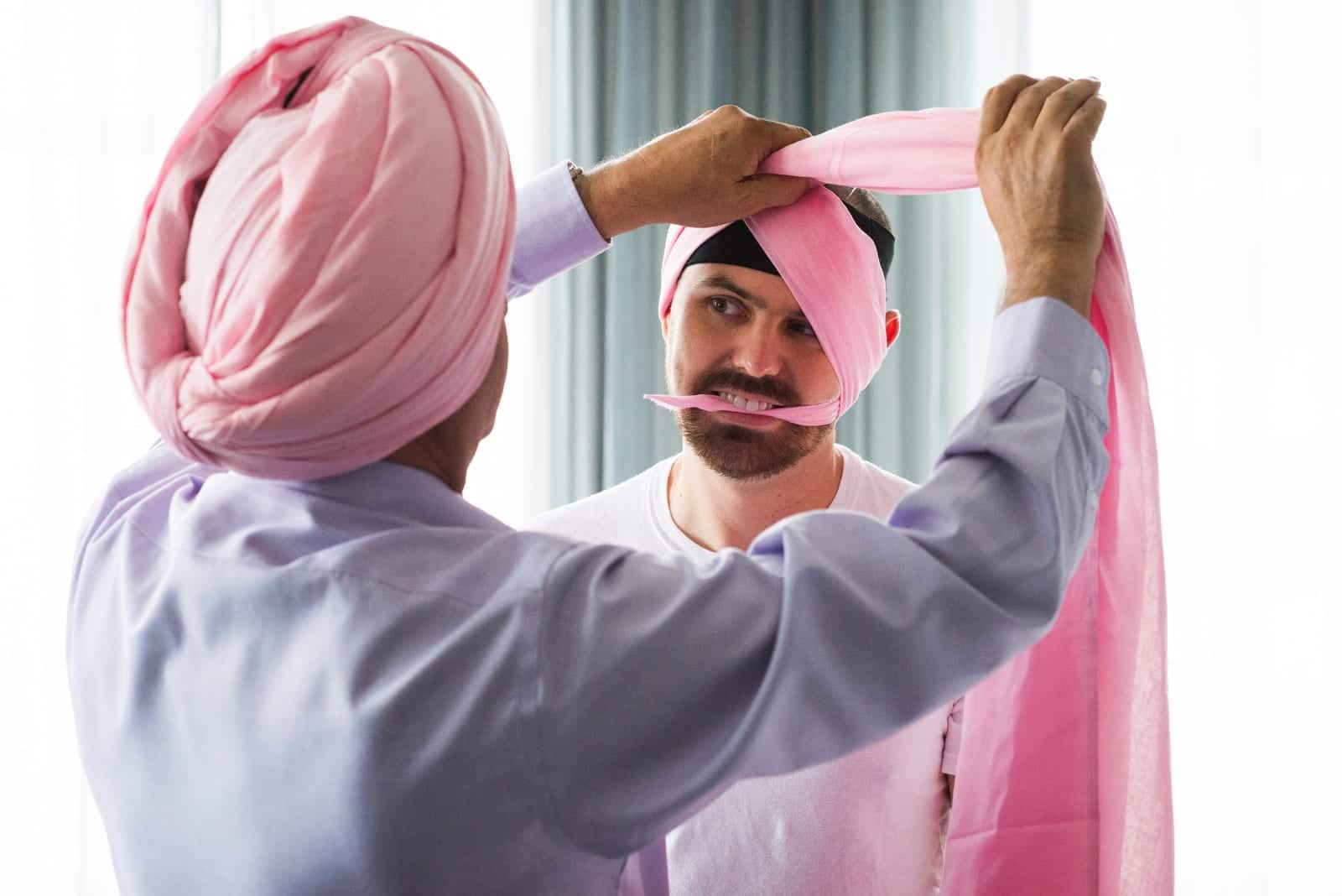 A groom holds one end of a pink cloth in his mouth as another man ties his turban before his South Asian wedding at the Renaissance Pittsburgh hotel.