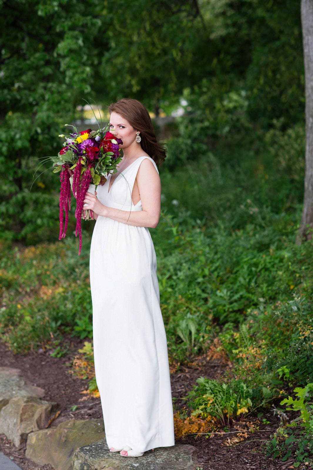 A bride holds her bouquet with pink, red, yellow and green flowers to her nose at Point State Park.