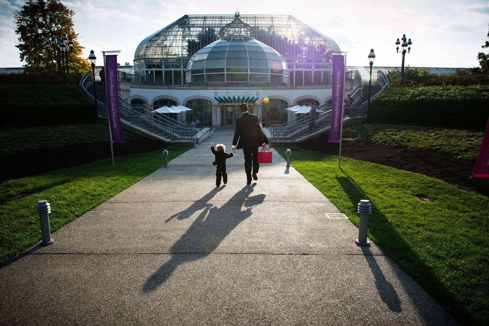 A groom carrying a red bag holds the hand of a small boy as they walk to Phipps Conservatory before his wedding. The setting sun casts long shadows on the path behind them.