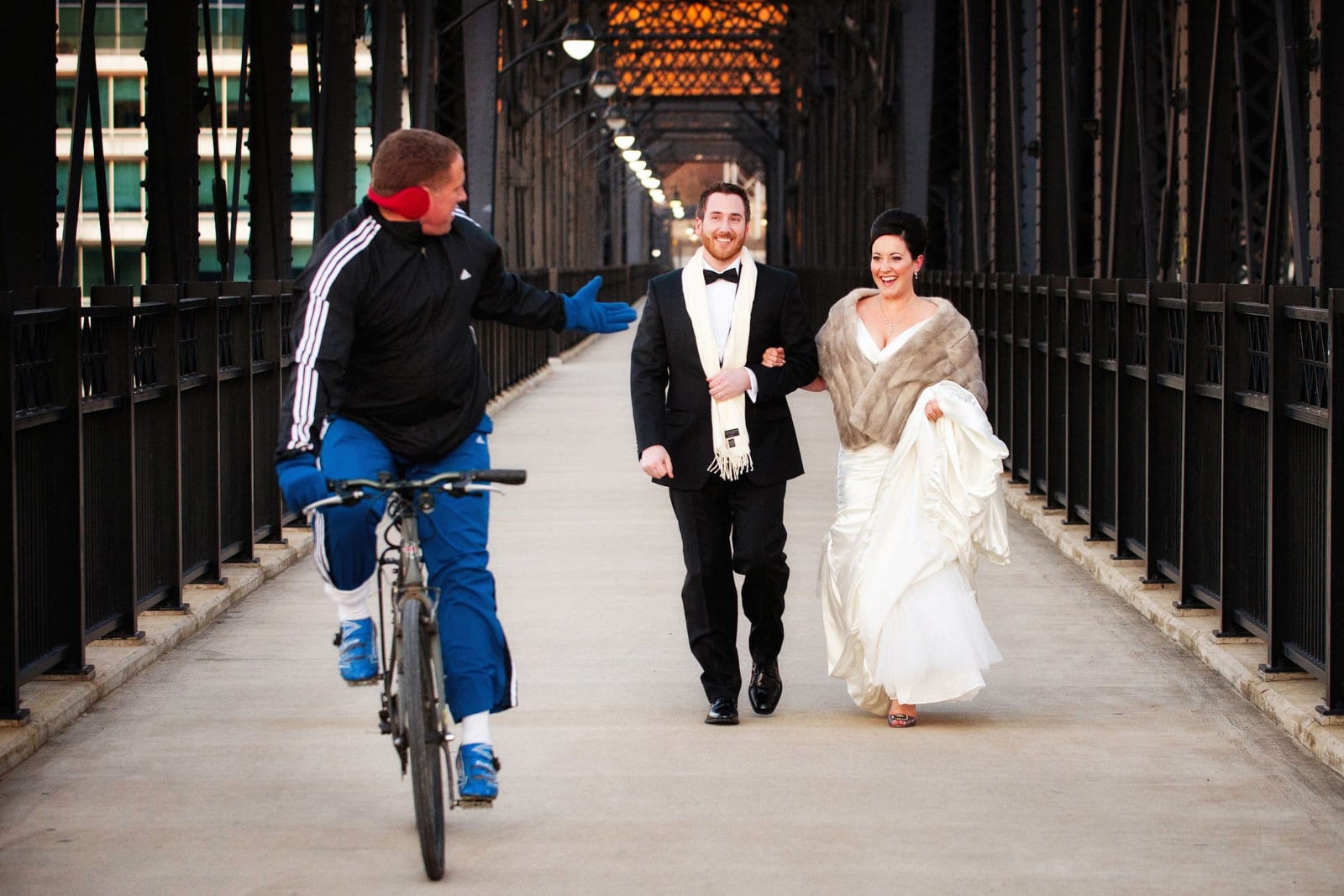 A man on a bicycle gestures to a bride and groom wearing a white scarf and a fur stole walk across the Hot Metal Bridge before their Andy Warhol Museum wedding.