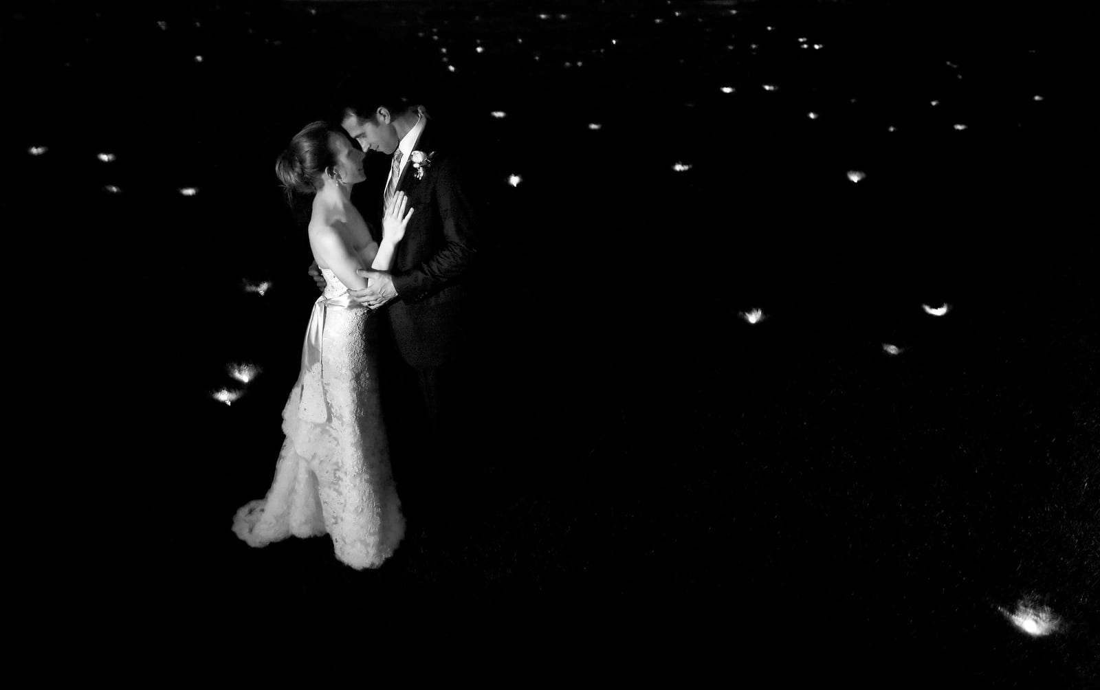 A bride and groom stand face to face in a dark field of lights.