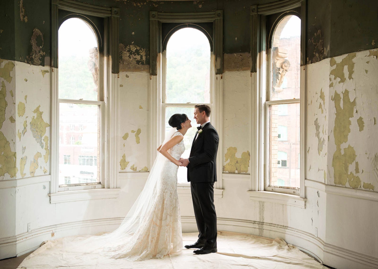 A bride and groom stand face-to-face in front of three tall arched windows in an old room with peeling paint.