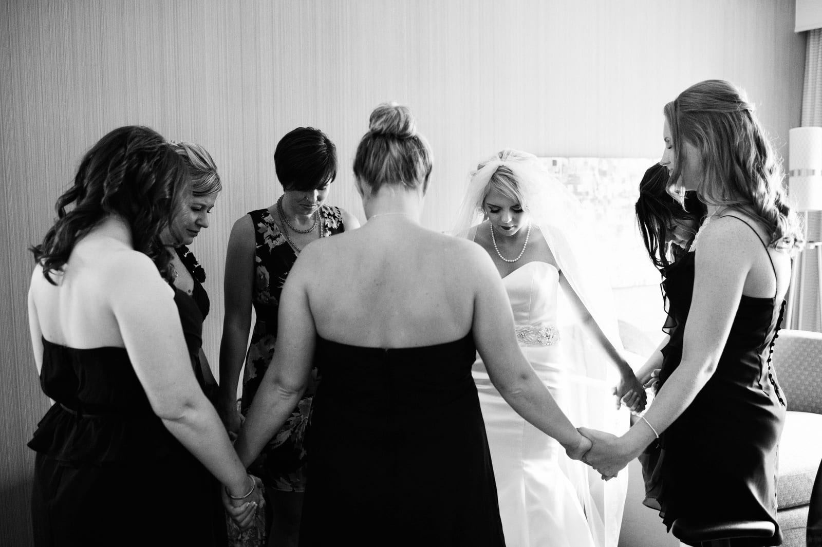 A bride and her bridesmaids hold hands and stand in a circle as they pray in a hotel room before the wedding.