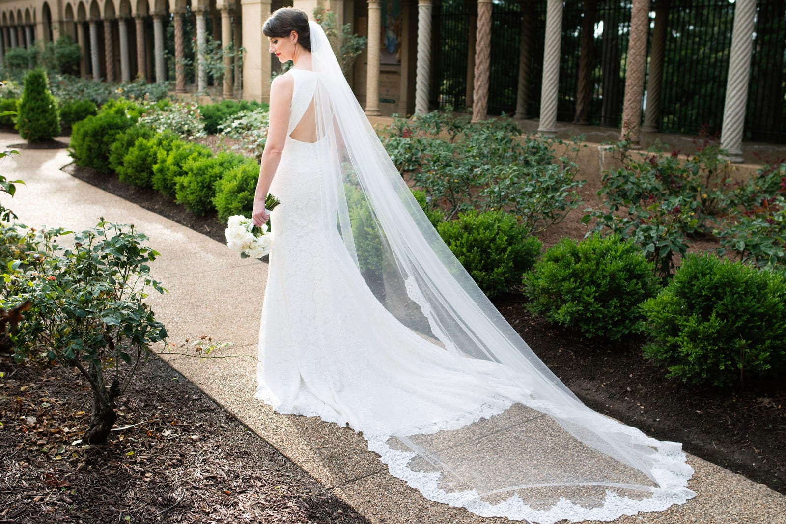 A bride looks back over her shoulder as she stands on a path with her veil and dress train spread out behind her.