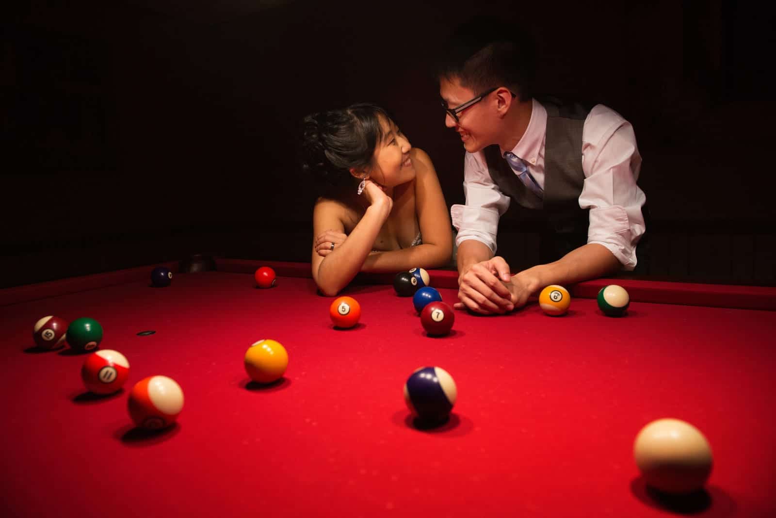 A bride and groom smile at each other as they lean on a pool table with a red felt top at the Grand Estate at Hidden Acres.