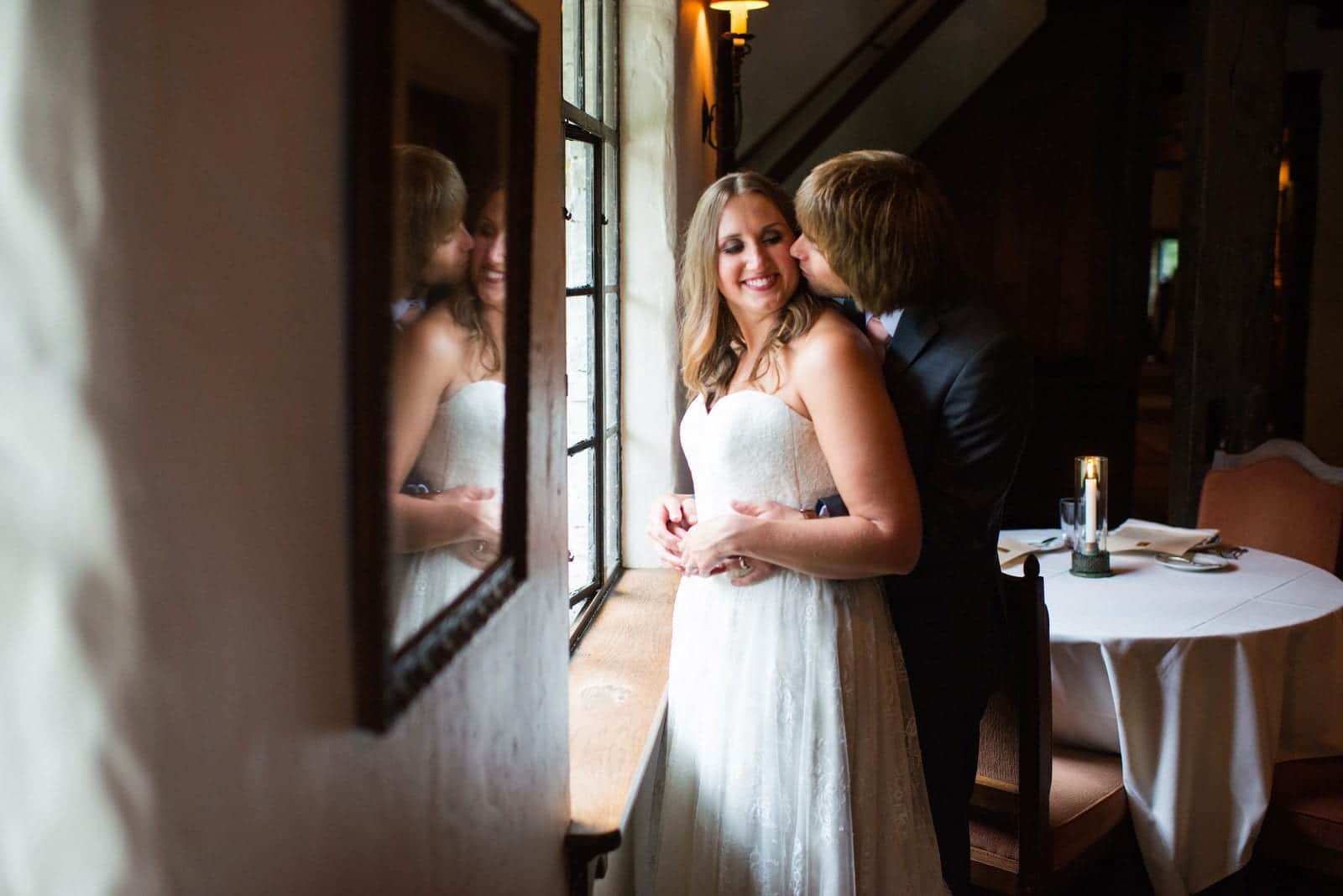 A bride smiles as her groom kisses her cheek in a window during their wedding at the Hyeholde in Moon Township.