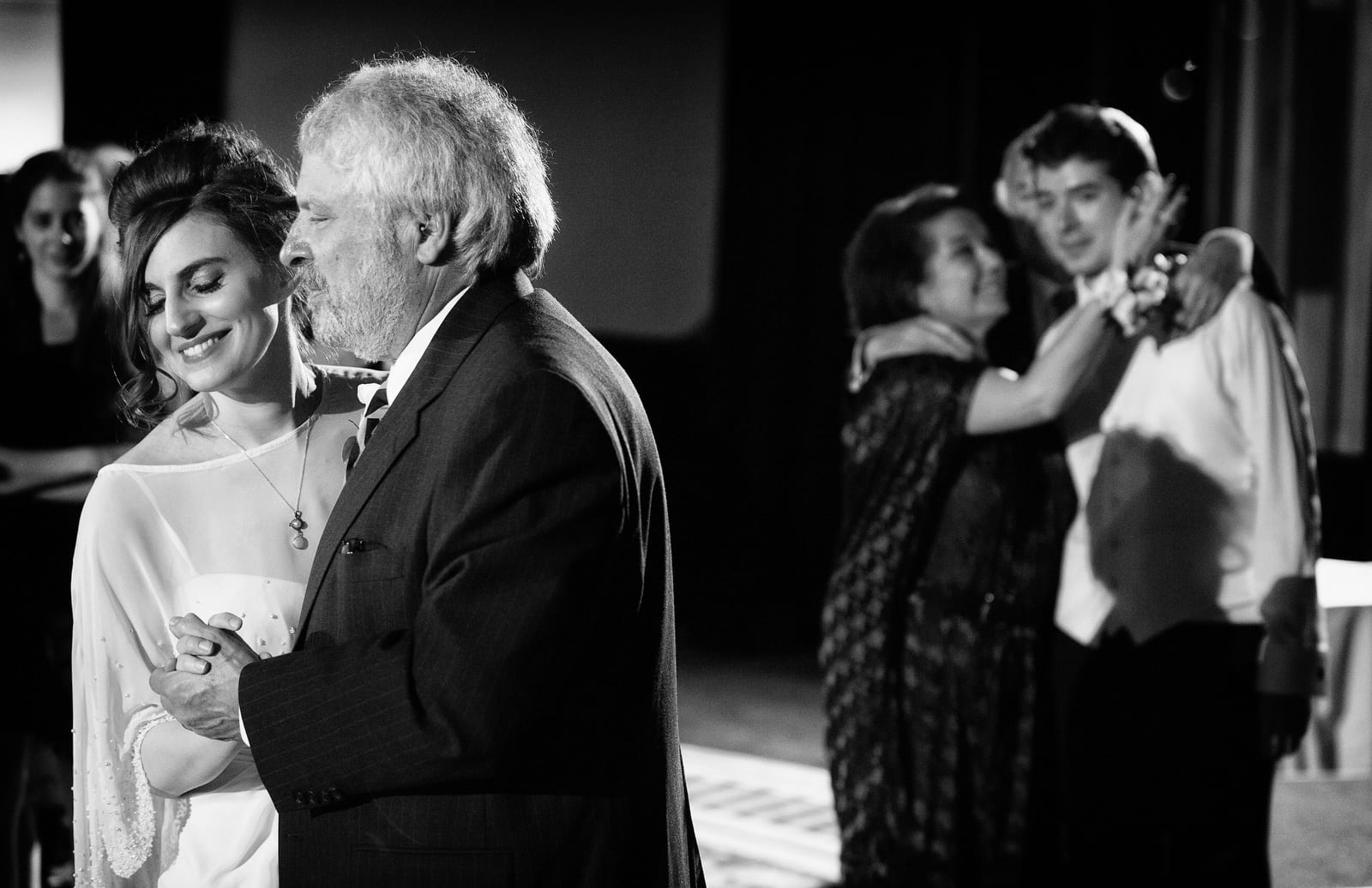 A bride and her father dance while the groom watches in the background as his happy parents embrace him.