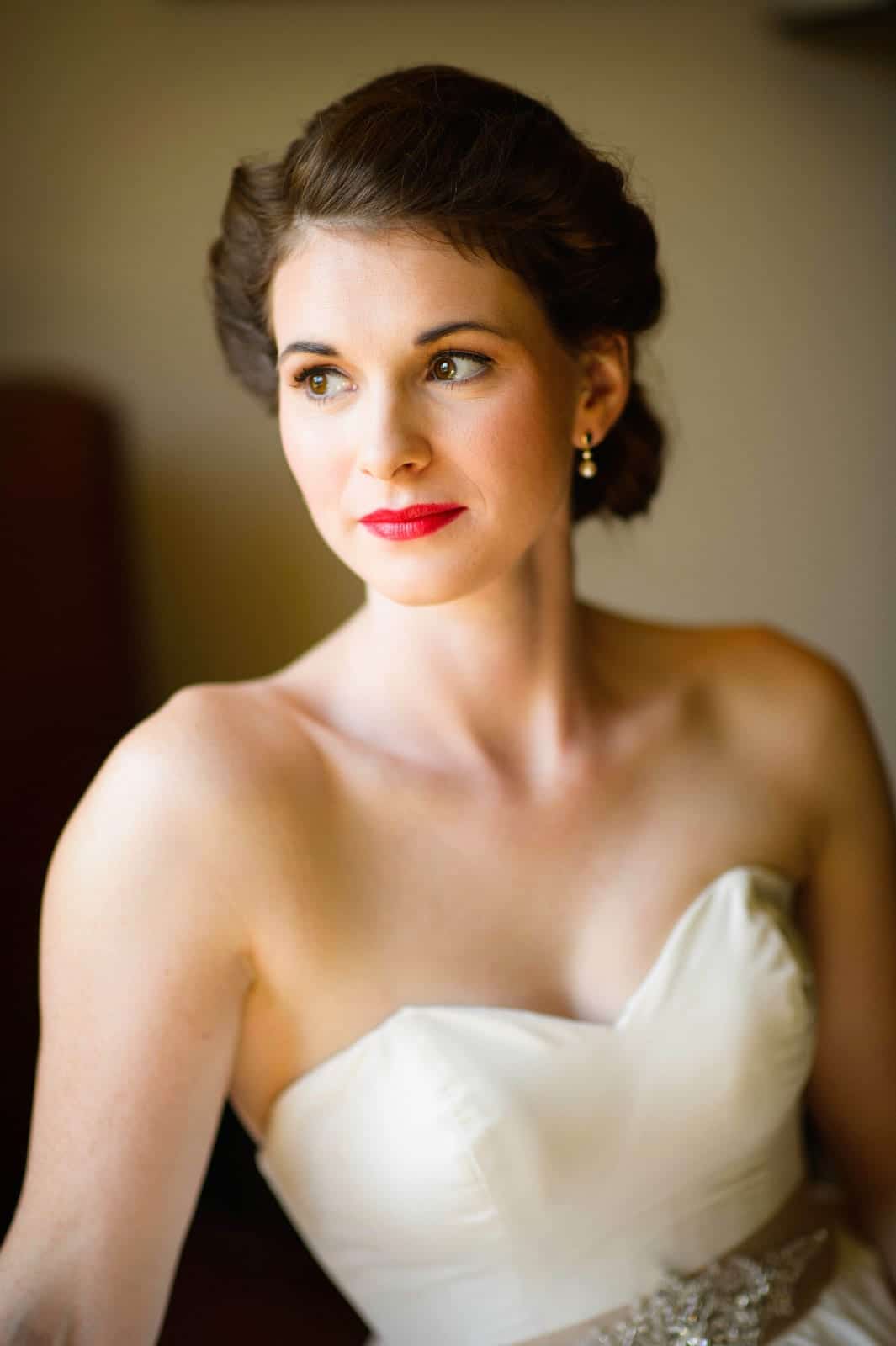 A brunette bride wearing bright red lipstick in a strapless wedding gown looks towards the left side of the frame.