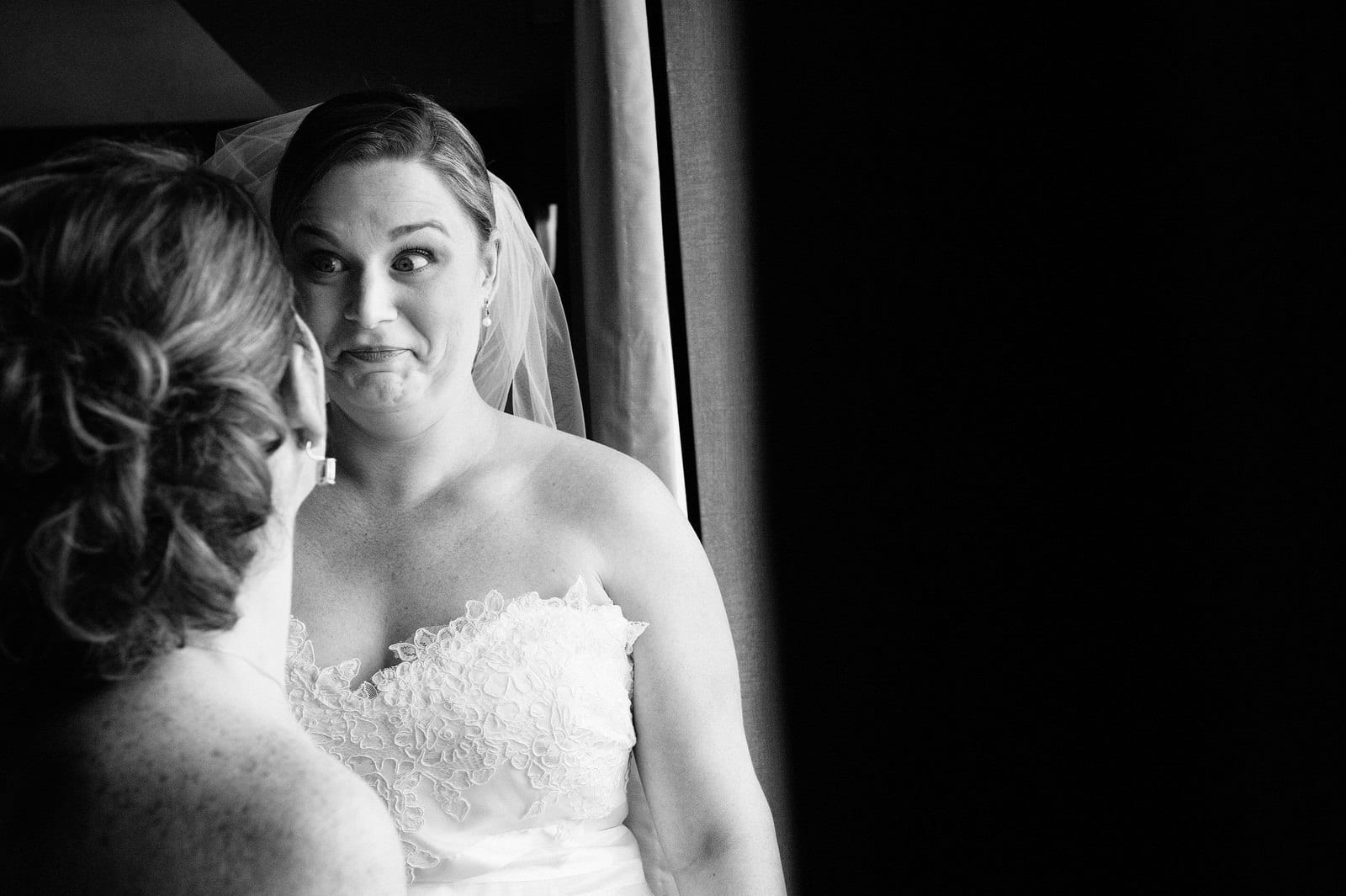 A bride makes a funny face to her bridesmaid after looking out the window of her hotel room.