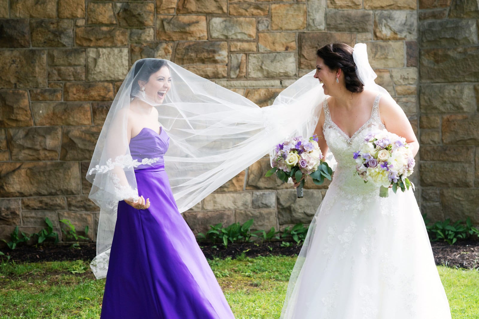 A bride laughs with her sister who wears a purple dress while standing under the veil before the bride's wedding at the Grand Concourse.