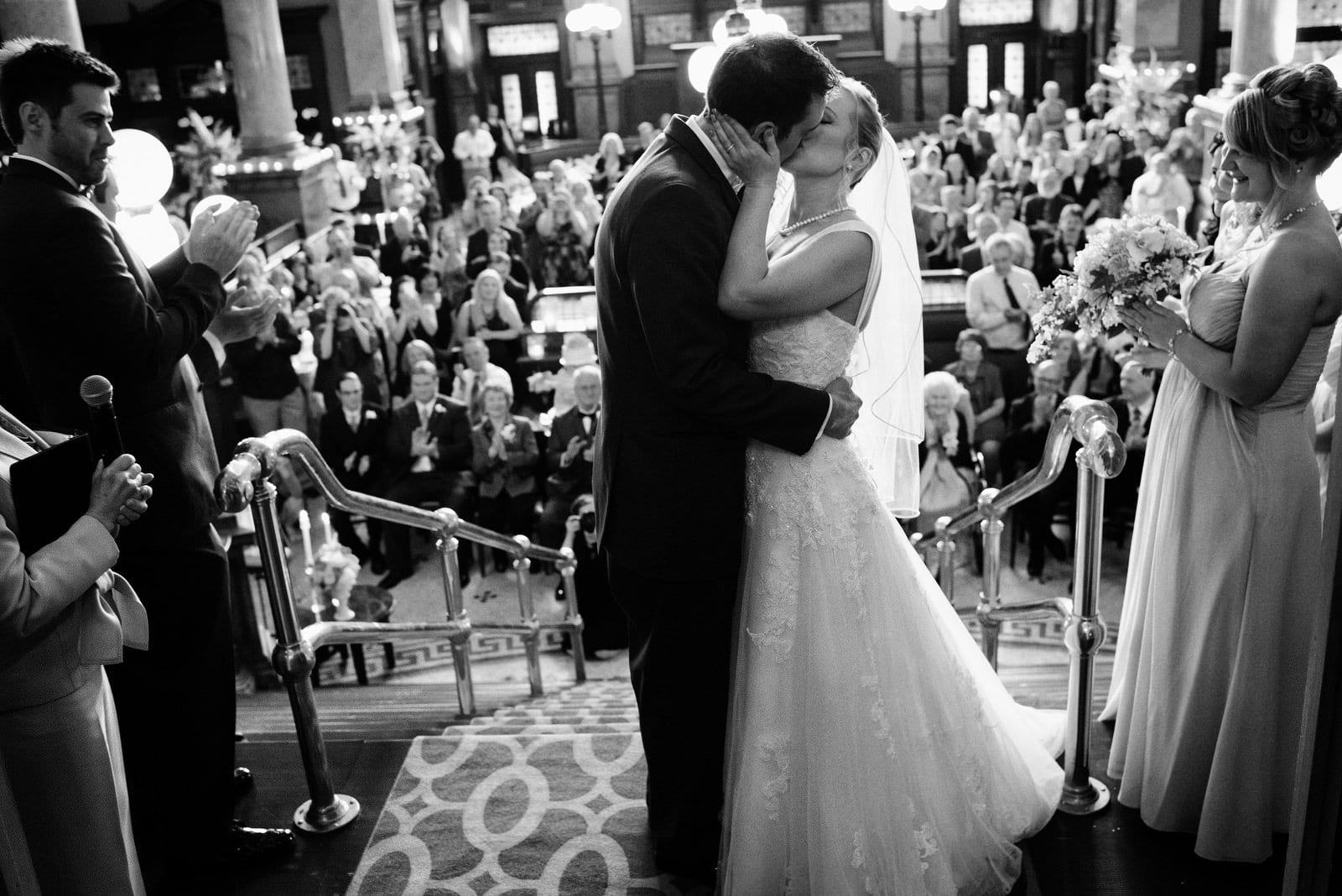 A bride and groom kiss on the landing of the Grand Concourse at the end of their wedding ceremony as their guests and bridal party behind them applaud.