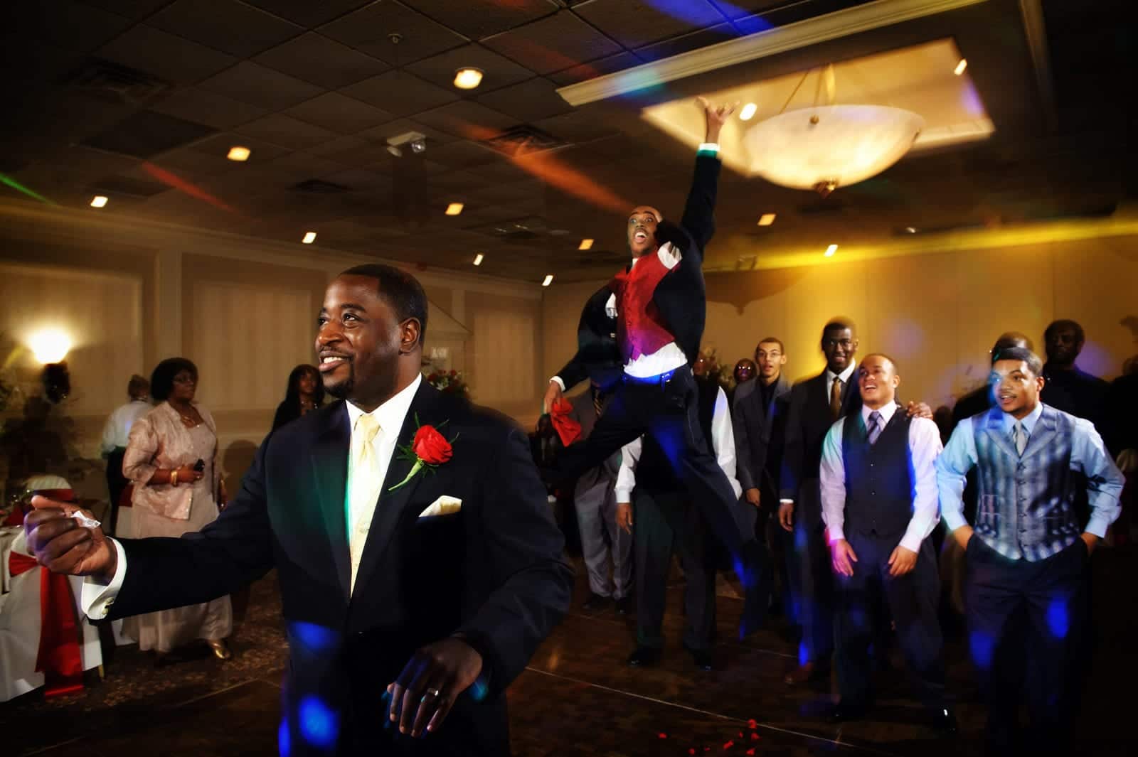 A groom smiles as he stands with his back to a group of men while preparing to throw a garter belt. There is a man jumping high into the air behind him.