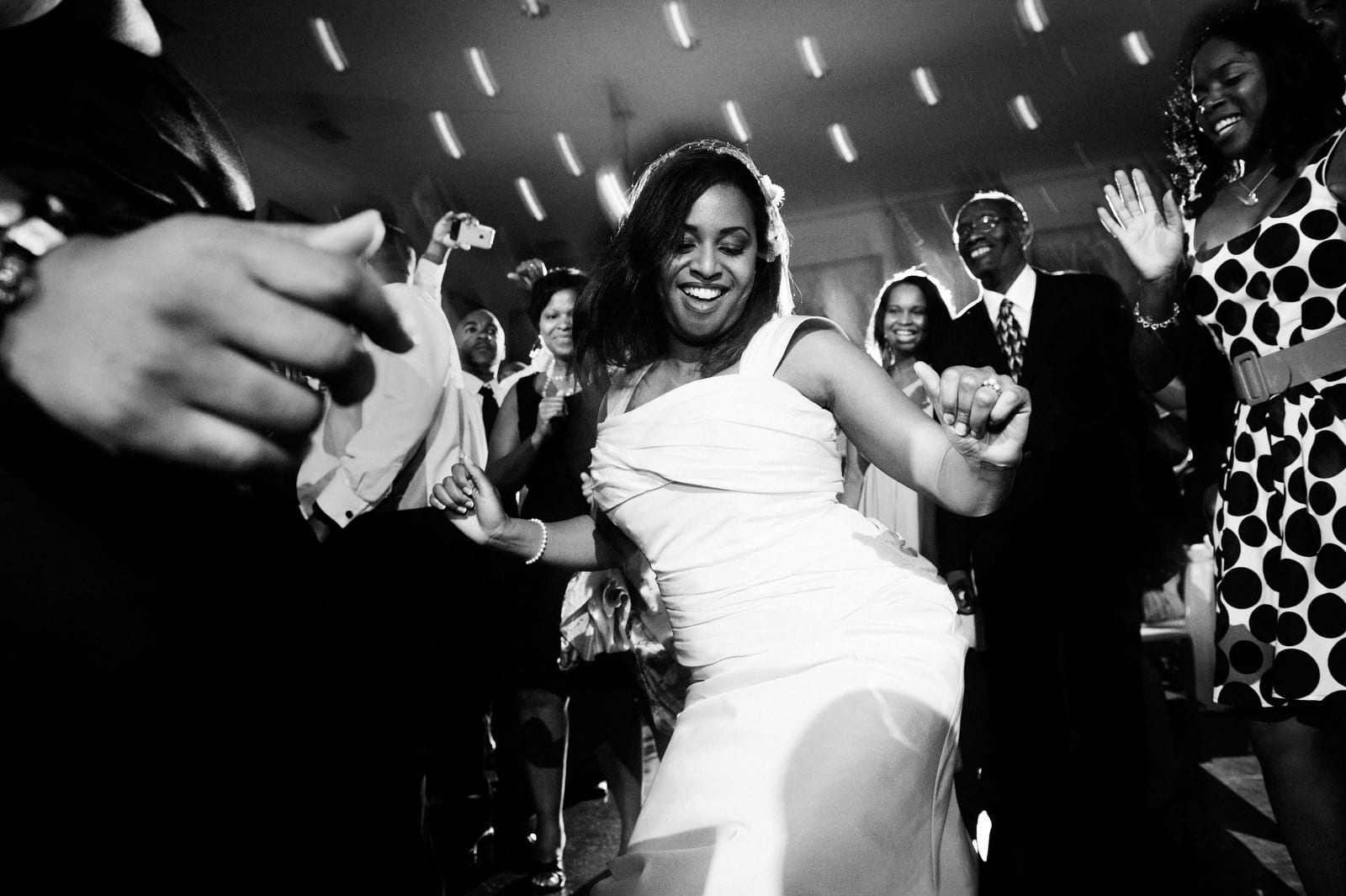 A bride in a white dress dances amongst a crowd of her guests. The small lights on the ceiling are streaks.