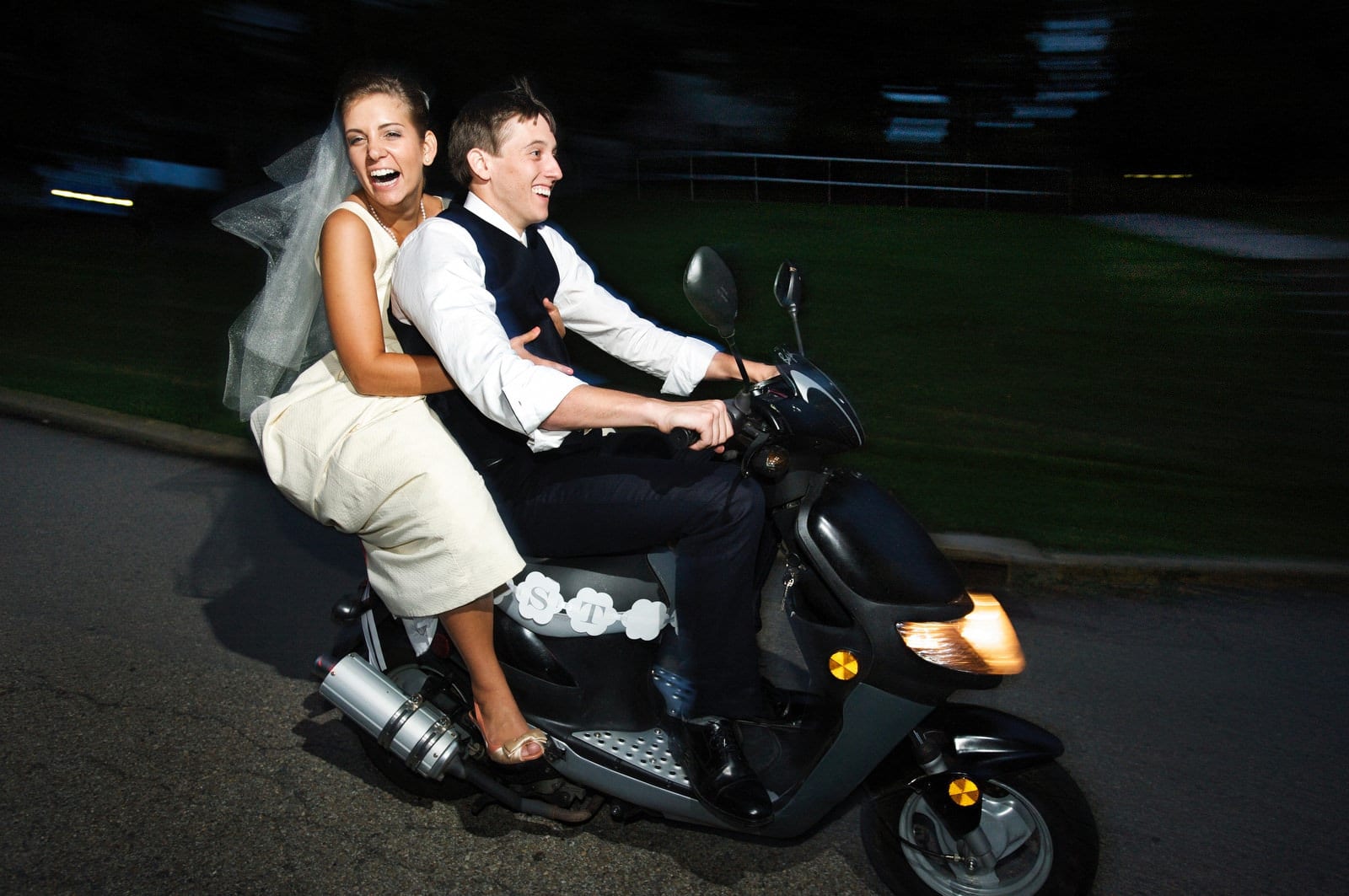 A bride laughs as she rides on the back of a motor scooter that her groom is driving.
