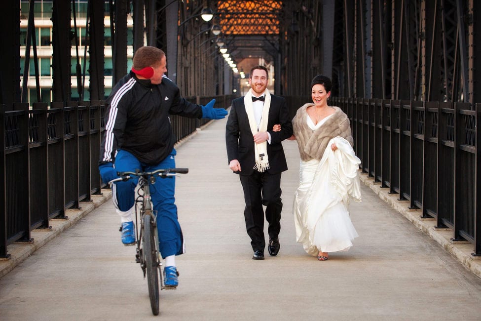 A bride and groom smile at a passing bicyclist as they walk across a bridge arm in arm