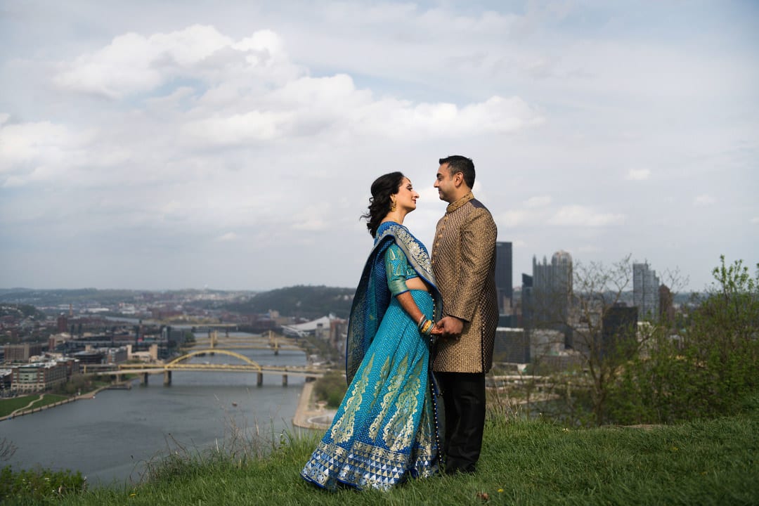 A couple wearing traditional Indian wedding garb stands on a hilltop with a view of downtown Pittsburgh behind them before their Indian Wedding Pittsburgh Wyndham.