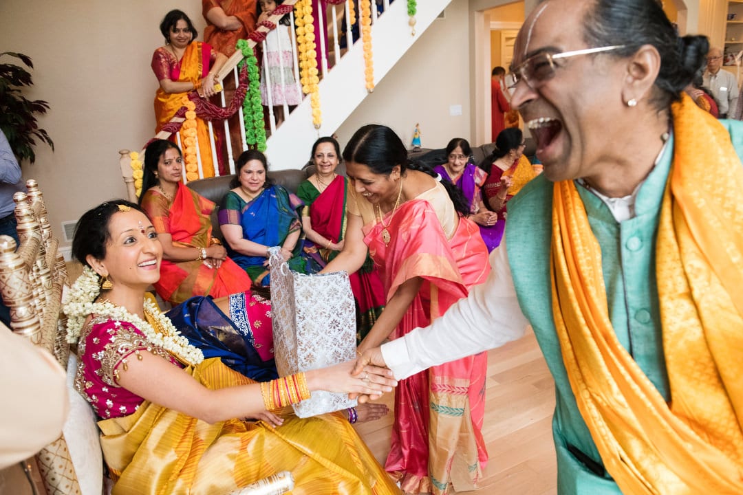 An Indian bride wearing a red and yellow sari shakes hands with a laughing Hindu priest.