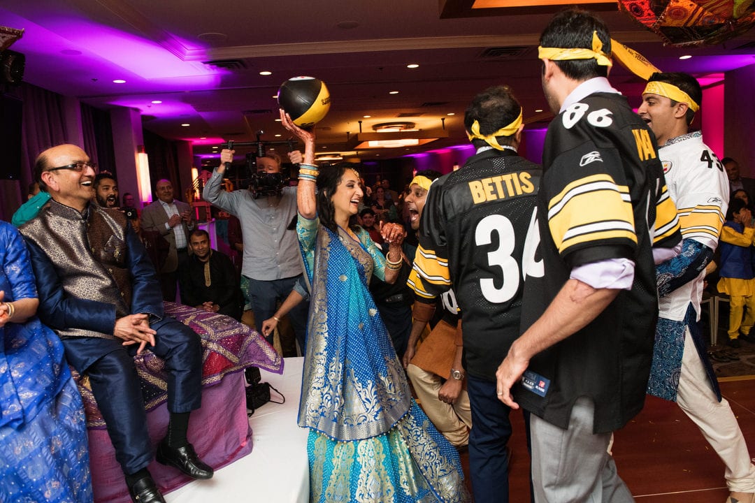 A bride laughs and holds a football above her head as wedding guests in Pittsburgh Steelers gear surround her.