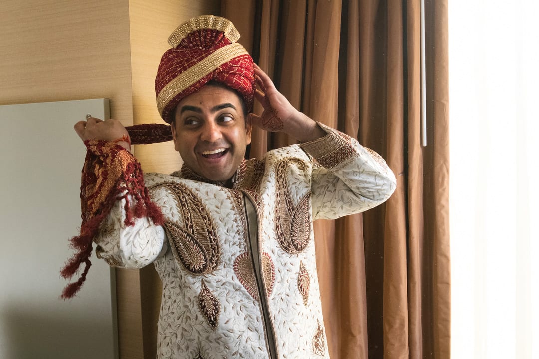 A groom smiles as he puts on a red and gold turban.