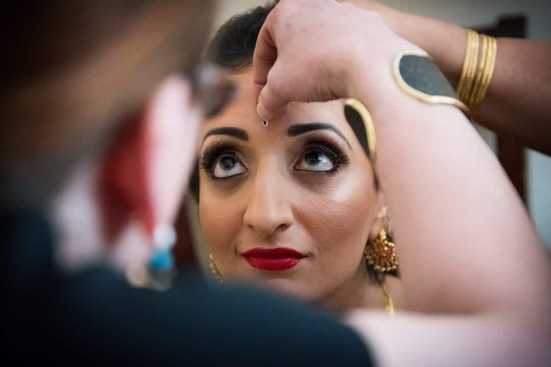 A bride looks up with her eyes as a makeup artist applies her bindi.