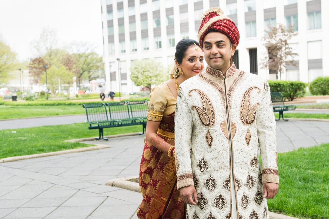 A bride in a red and gold sari sneaks up on her groom wearing a white and gold tunic and a red and gold turban during their first look.