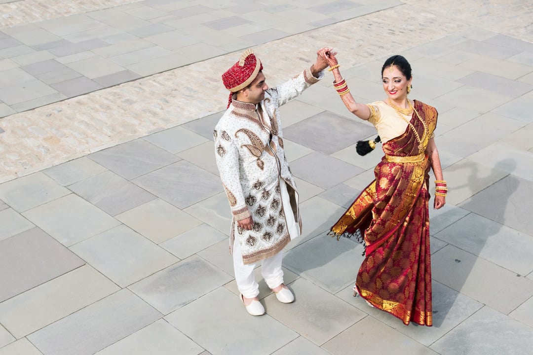 Seen from above, a groom wearing a gold and white tunic spins his bride in her red and gold sari.