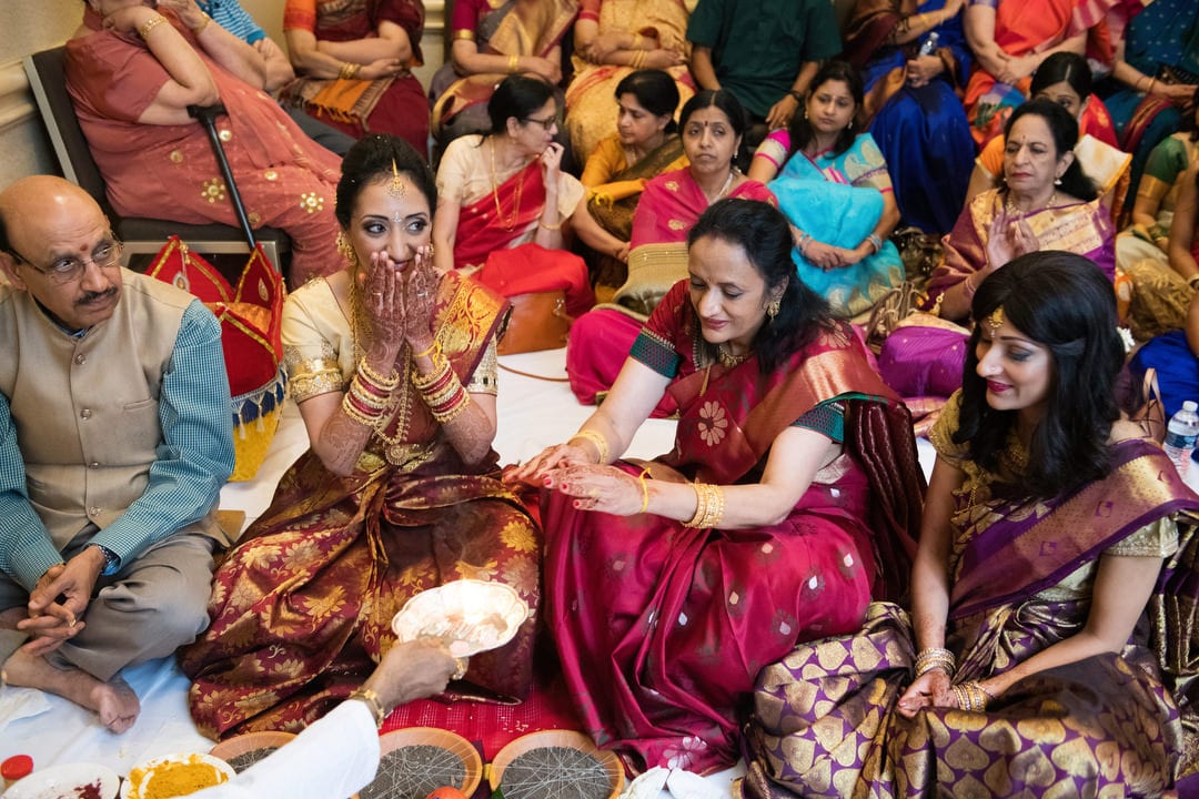 Indian men and women sit on the floor of a room during a Gauri Puja.